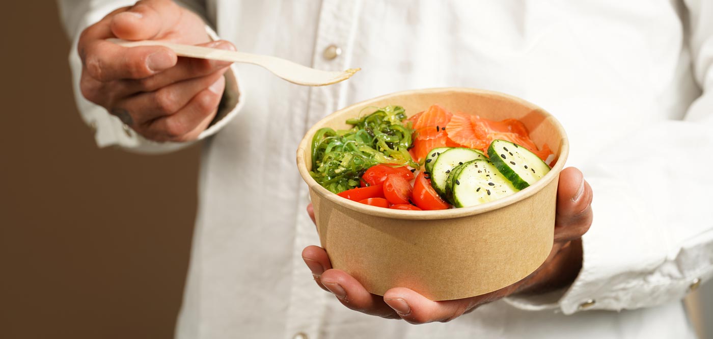 Mann hält eine Poke-Schüssel – Reis, Wakame-Algen, Tomaten, Gurken, Avocado, Lachs in recycelter Verpackung mit nachhaltiger Gabel, Essen zum Mitnehmen